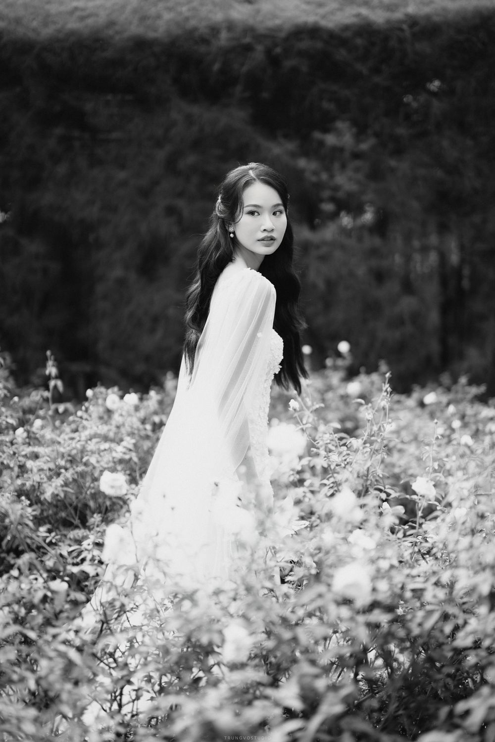 Young woman in white dress standing gracefully among blooming flowers in a garden, black and white photography.