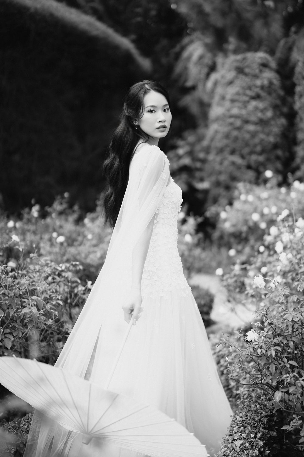 Elegant woman in flowing white gown holding umbrella among lush garden greenery, black and white portrait.