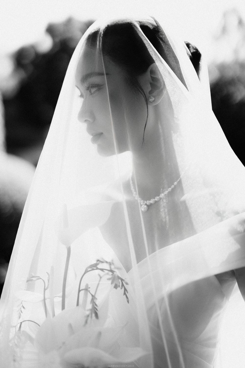 Beautiful bride wearing pearl necklace and sheer veil holding bouquet, soft black and white profile portrait.