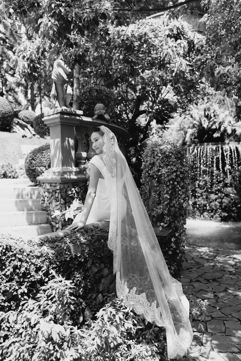Elegant bride in fitted wedding dress with long lace veil leaning on stone wall in garden setting.