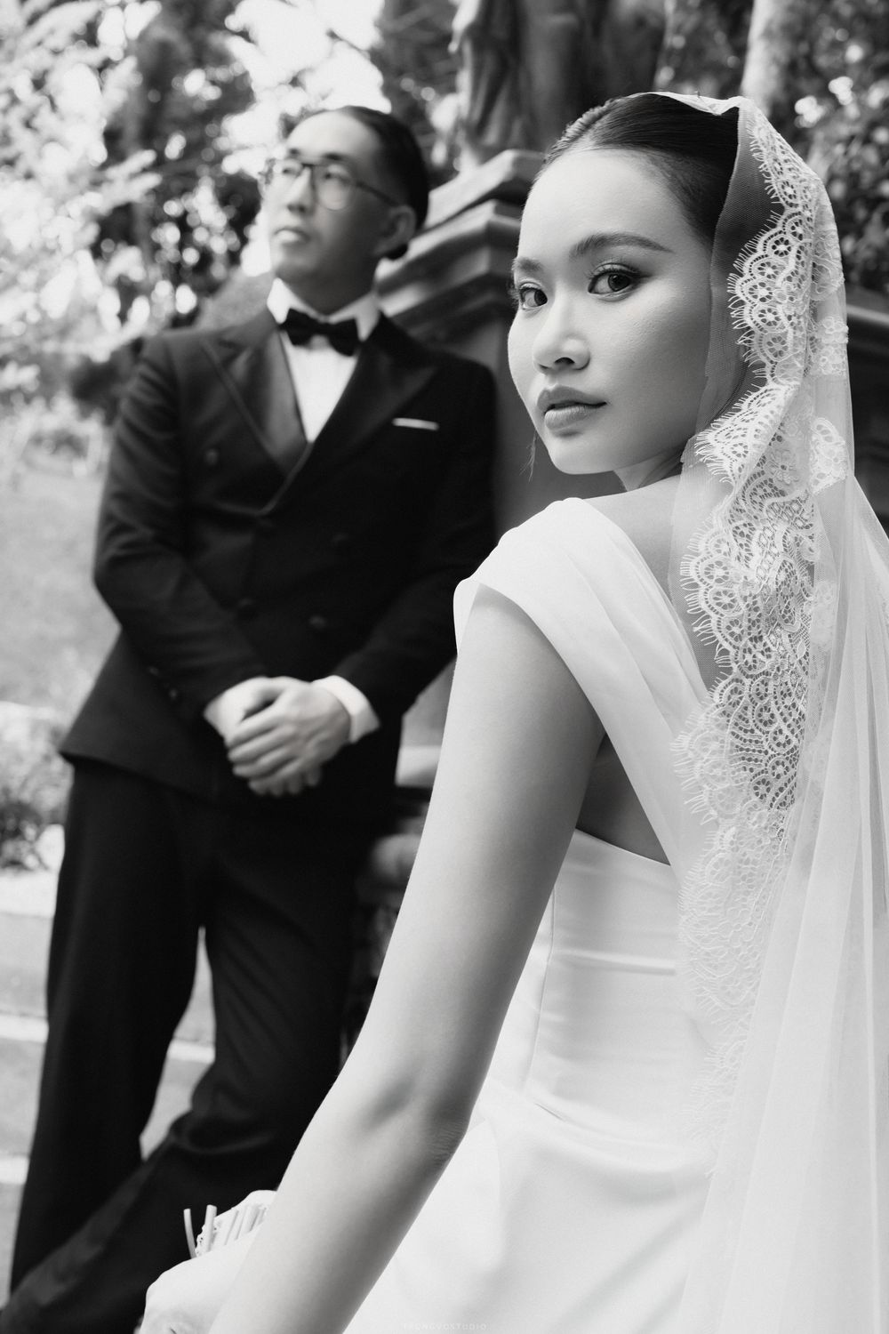 Elegant bride in white gown with lace veil gazing forward as groom stands in background, black and white.