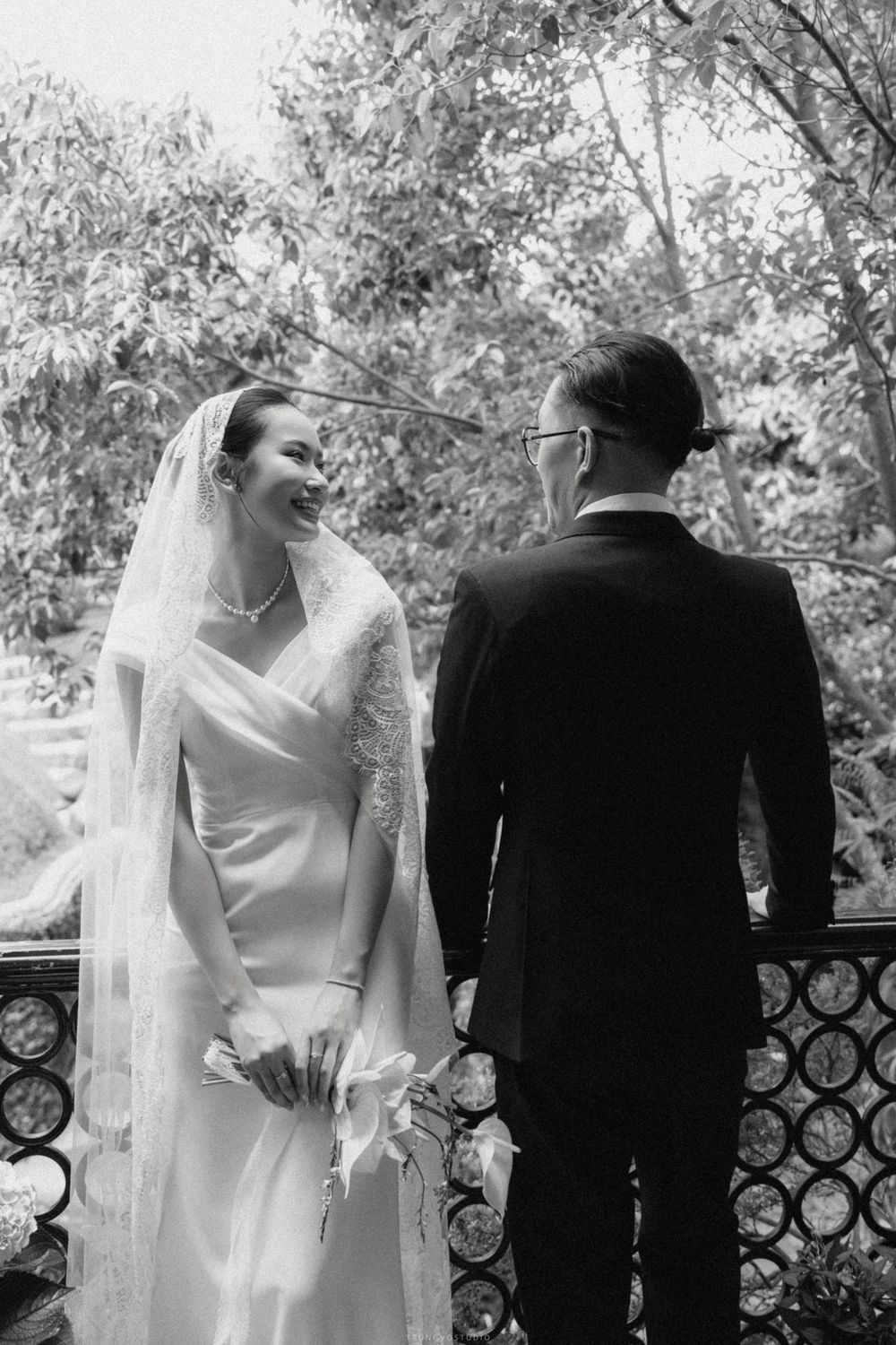Smiling bride in white dress with lace veil holding flowers while groom looks on, outdoor wedding portrait.