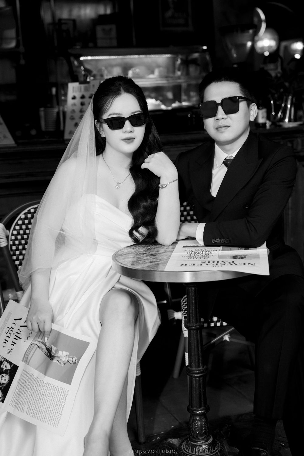 Black and white photo of a bride and groom wearing sunglasses sitting at a cafe table.
