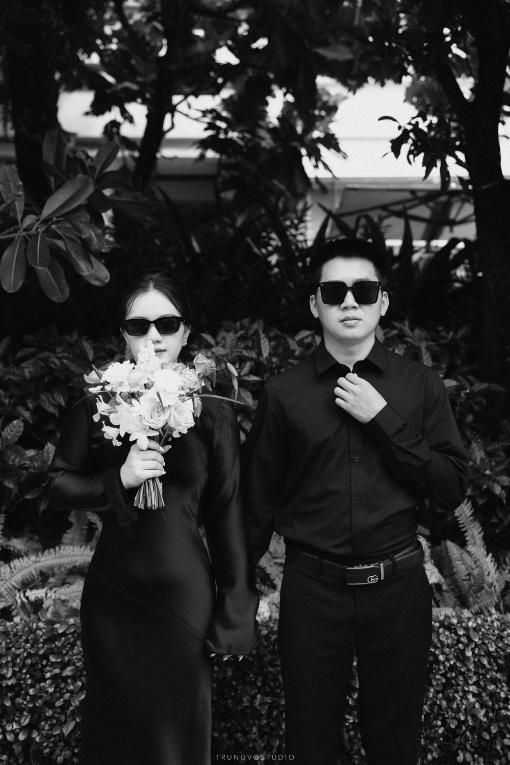 Black and white photo of a stylish couple in all-black outfits and sunglasses, woman holding white flowers.