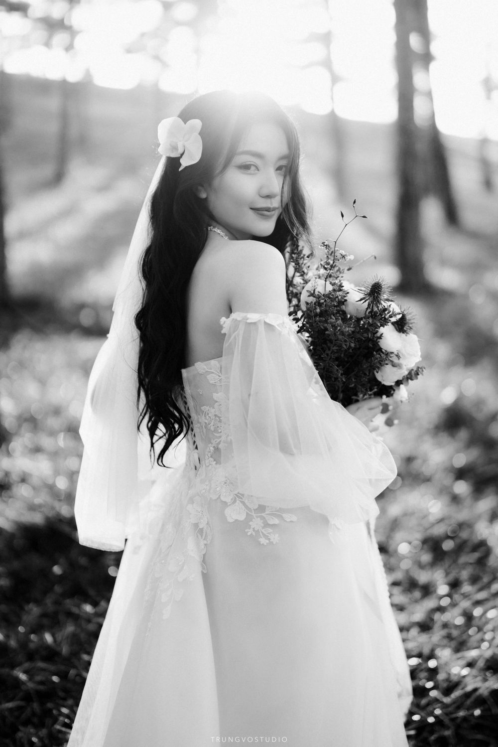 Elegant bride in white off-shoulder gown holding bouquet, photographed in sunlit forest in black and white.