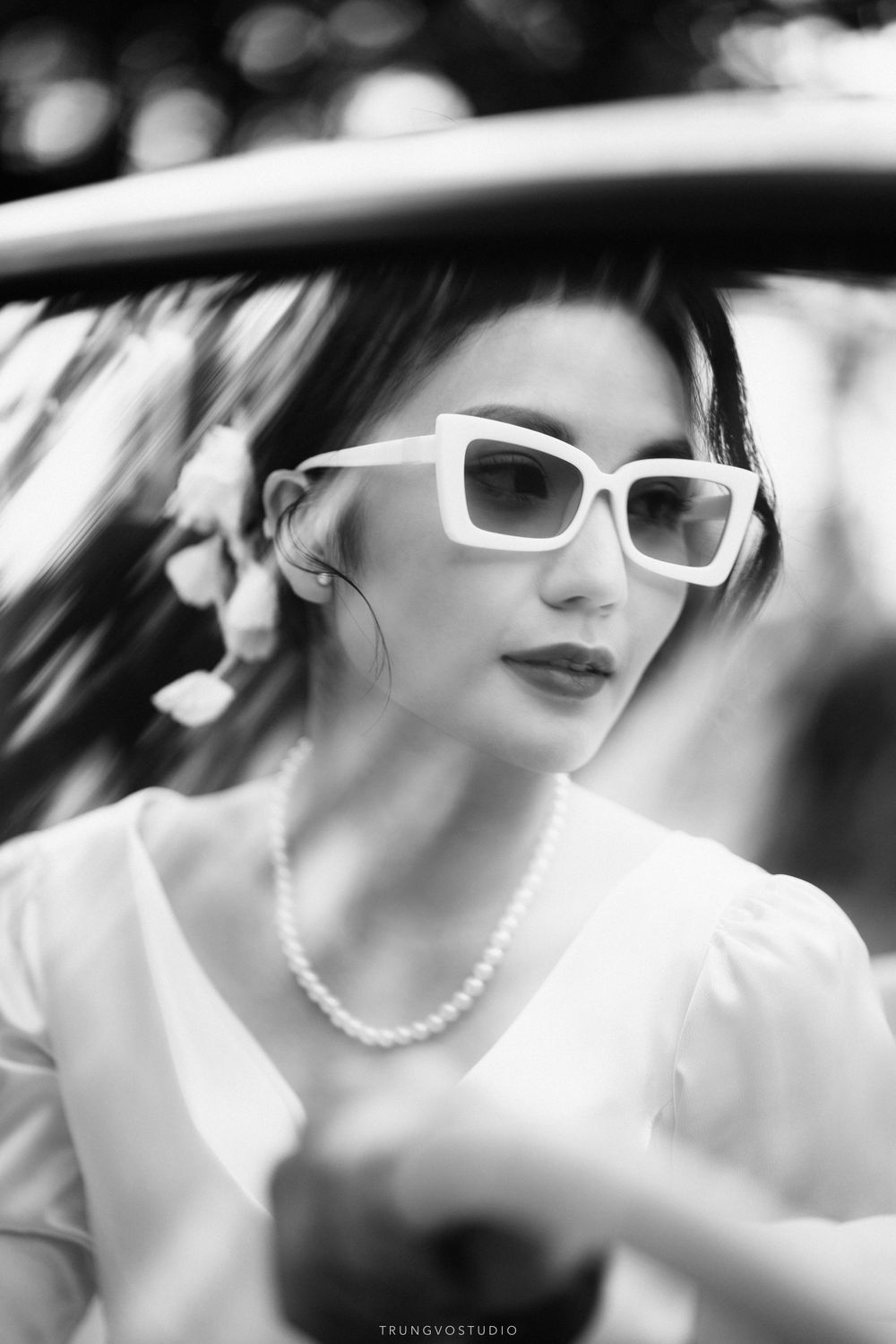 Black and white portrait of a chic woman in white sunglasses and pearl necklace sitting in a car.