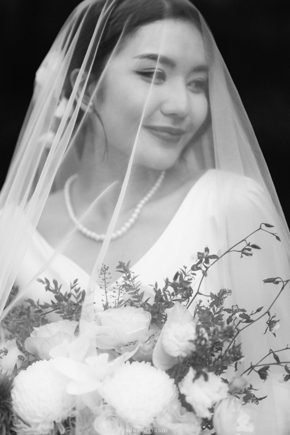 Dreamy black and white portrait of a veiled bride smiling softly while holding a lush floral bouquet.