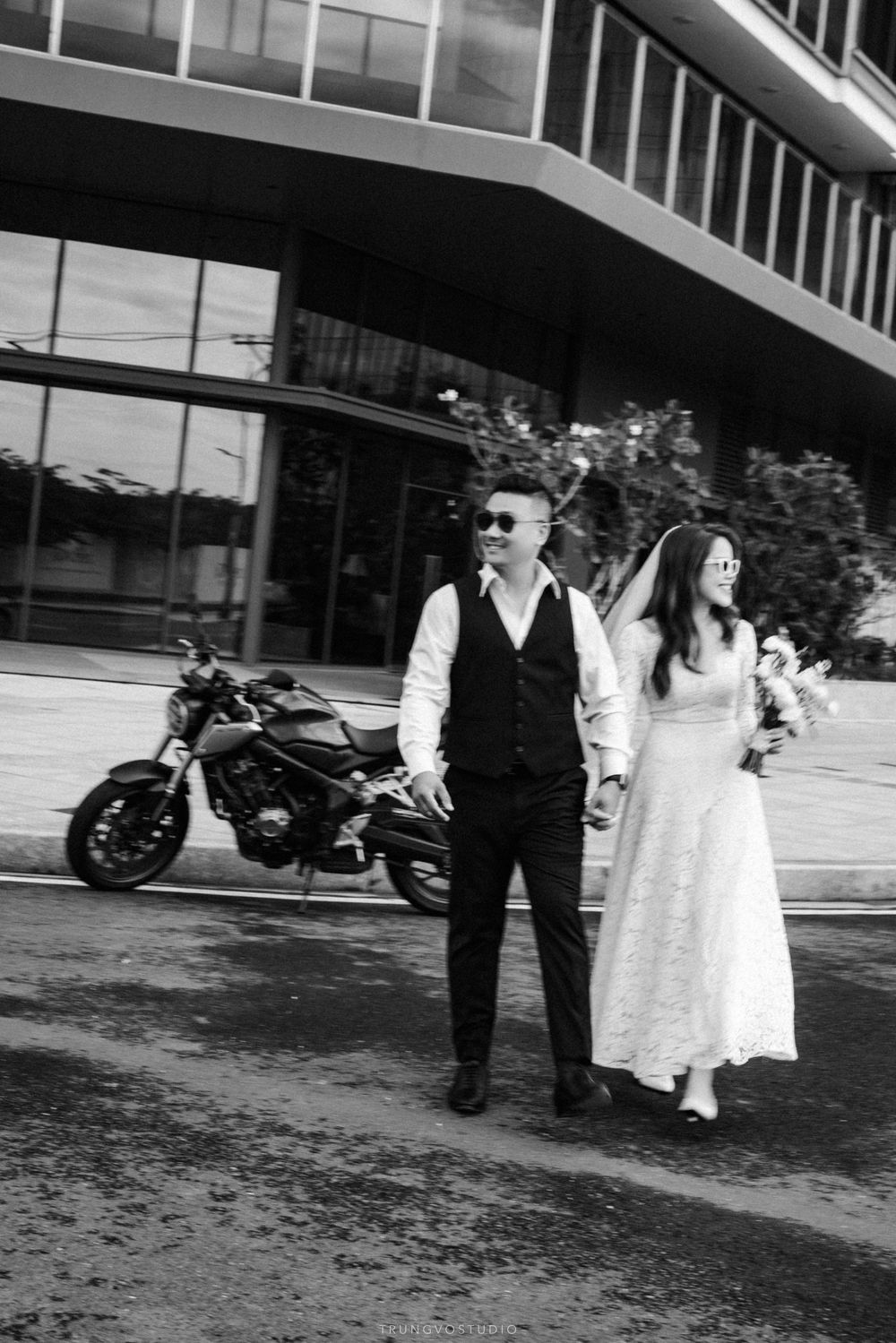 Black and white wedding photo of couple walking hand-in-hand with motorcycle and modern building backdrop.