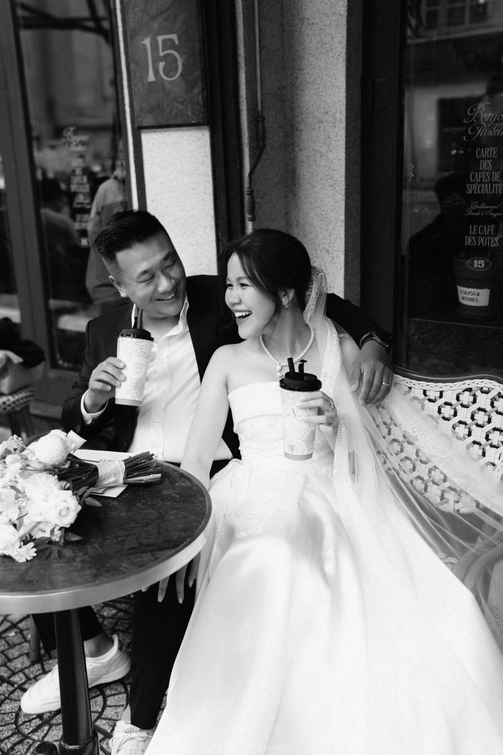 Black and white photo of a joyful wedding couple sharing drinks at a Parisian-style outdoor cafe.