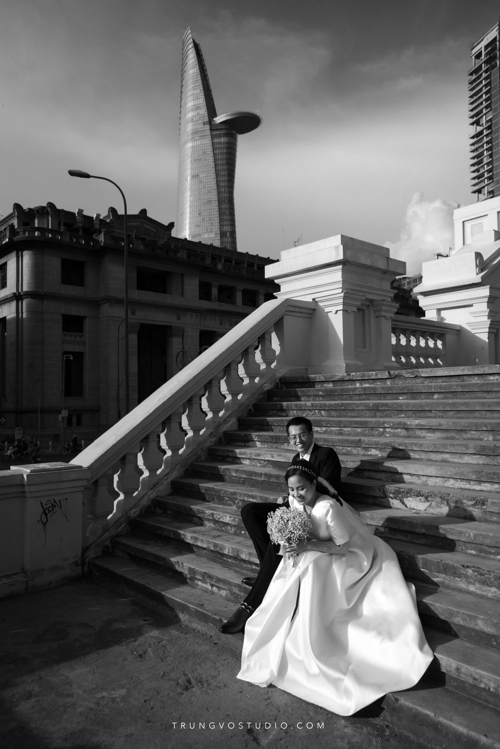 Dramatic black and white wedding photo of couple sitting on grand staircase with city landmark behind.