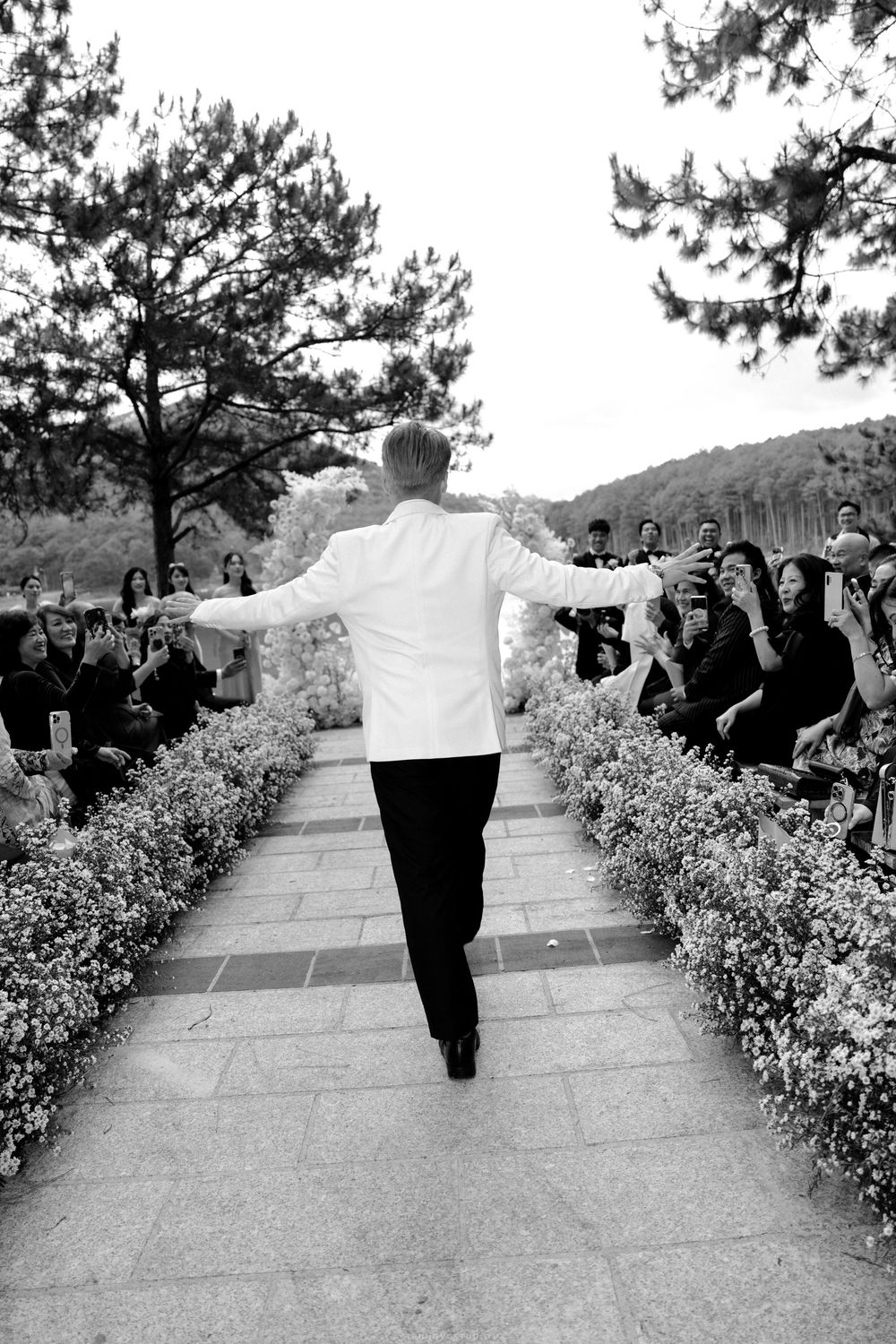 Black and white photo of a groom in white jacket walking down a flower-lined aisle toward guests.
