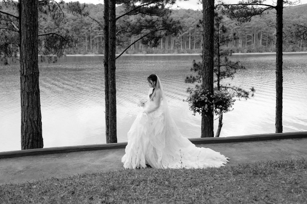 Bride in flowing white gown standing by lakeside among trees in black and white wedding portrait.