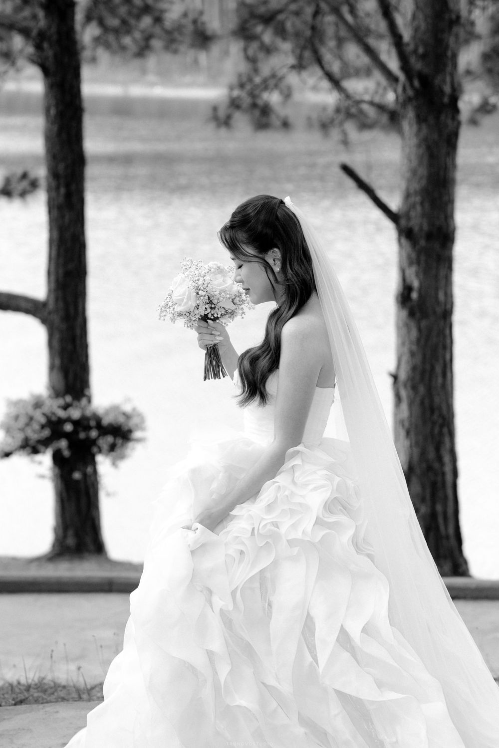 Black and white portrait of a bride in a ruffled gown smelling her bouquet near a lakeside.