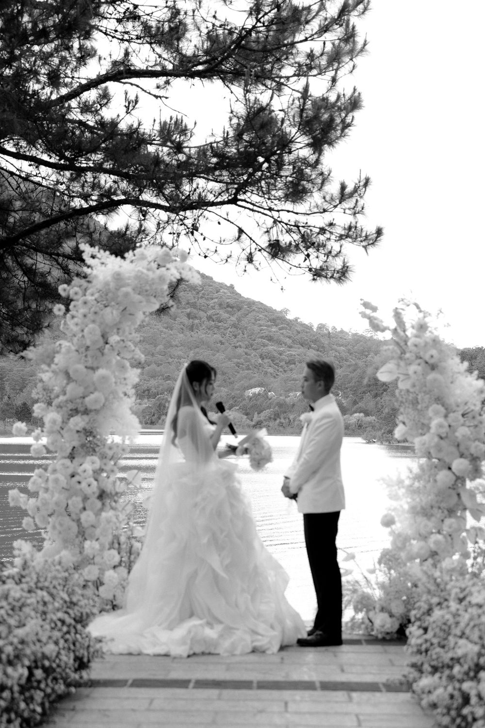 Black and white wedding ceremony photo of couple exchanging vows under an elegant floral arch.