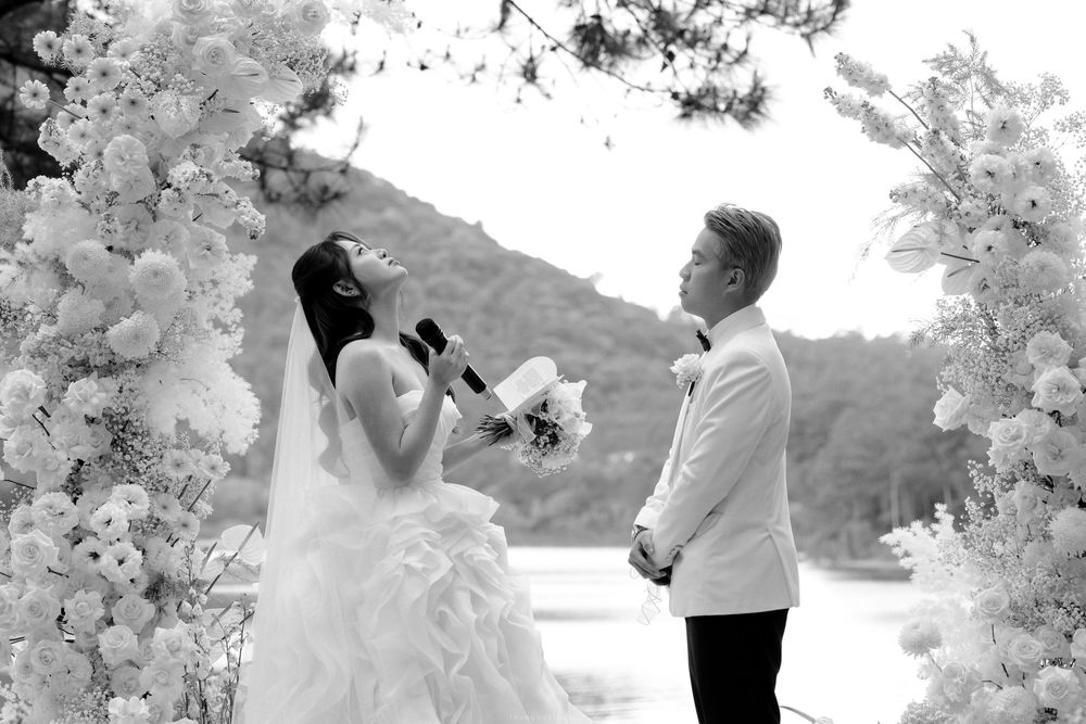 Black and white photo of a bride reading vows to her groom under a floral arch outdoors.