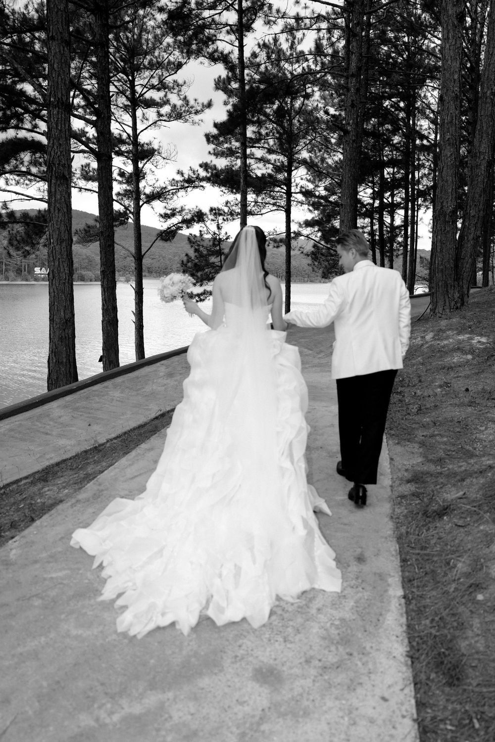 Black and white photo of bride and groom walking hand in hand along a lakeside path in pine trees.