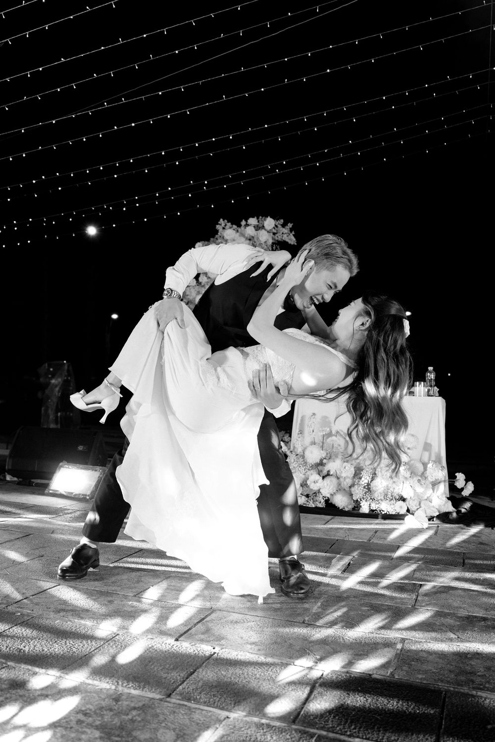 Black and white photo of groom dipping bride during first dance under string lights at wedding reception.