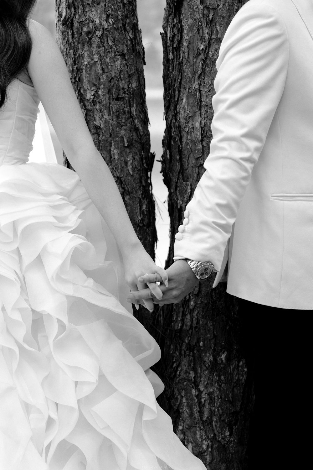 Black and white close-up of bride and groom holding hands in front of a tree trunk.