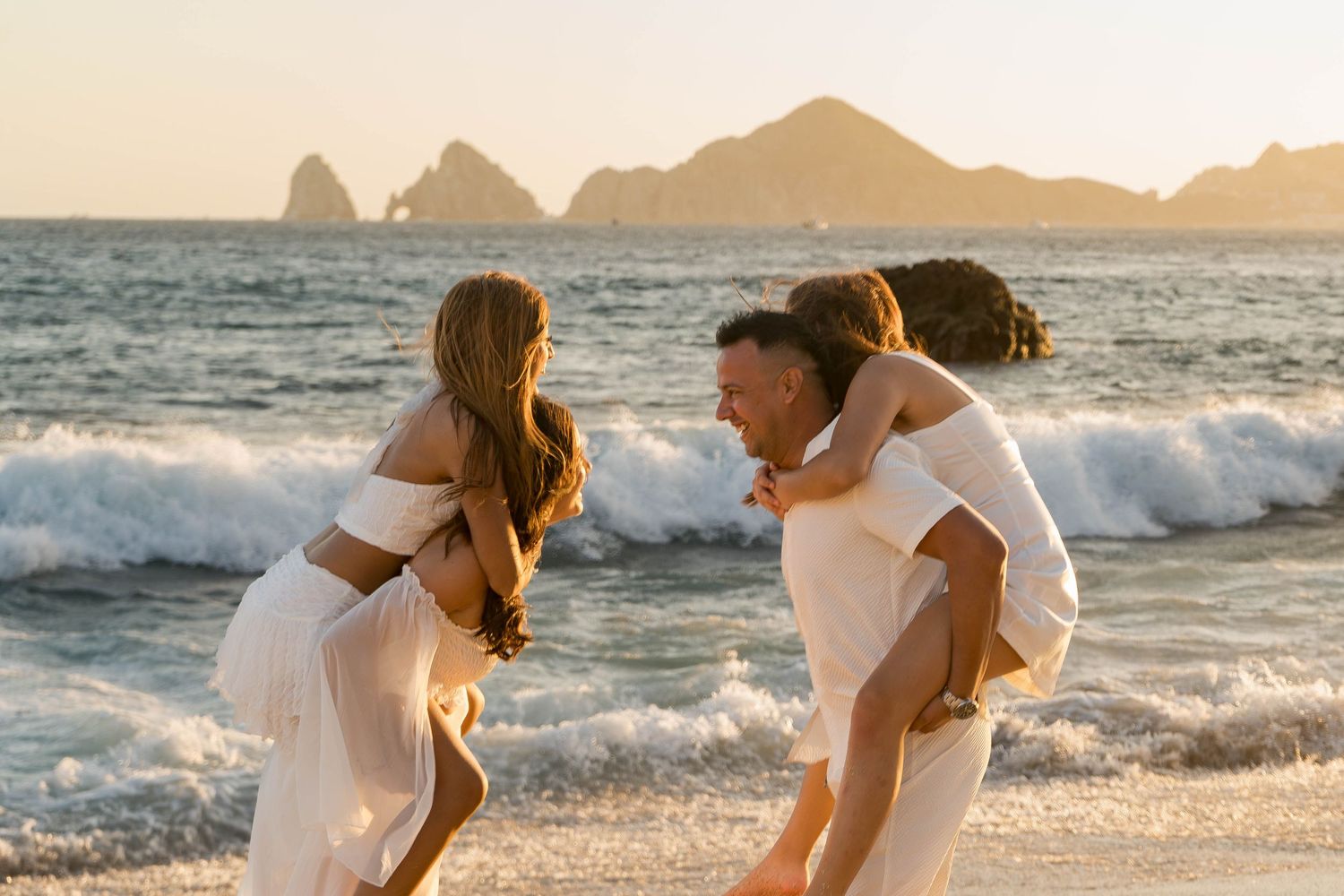 Couples enjoying a playful moment together on a beach at sunset with waves crashing behind them.