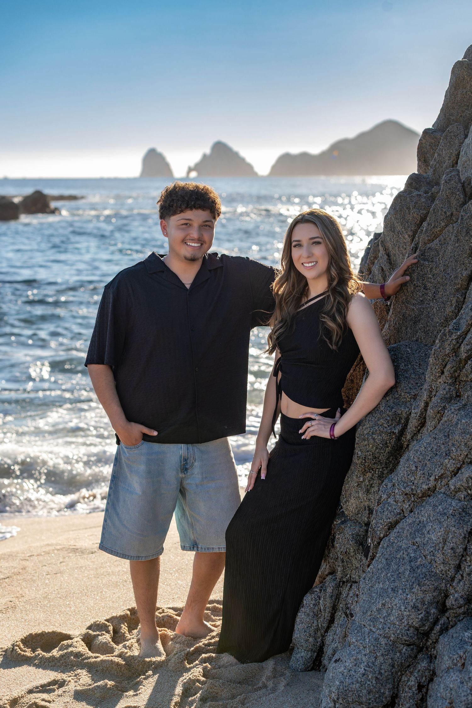 A couple poses together on a rocky beach with sparkling ocean waves and sunlight in the background.