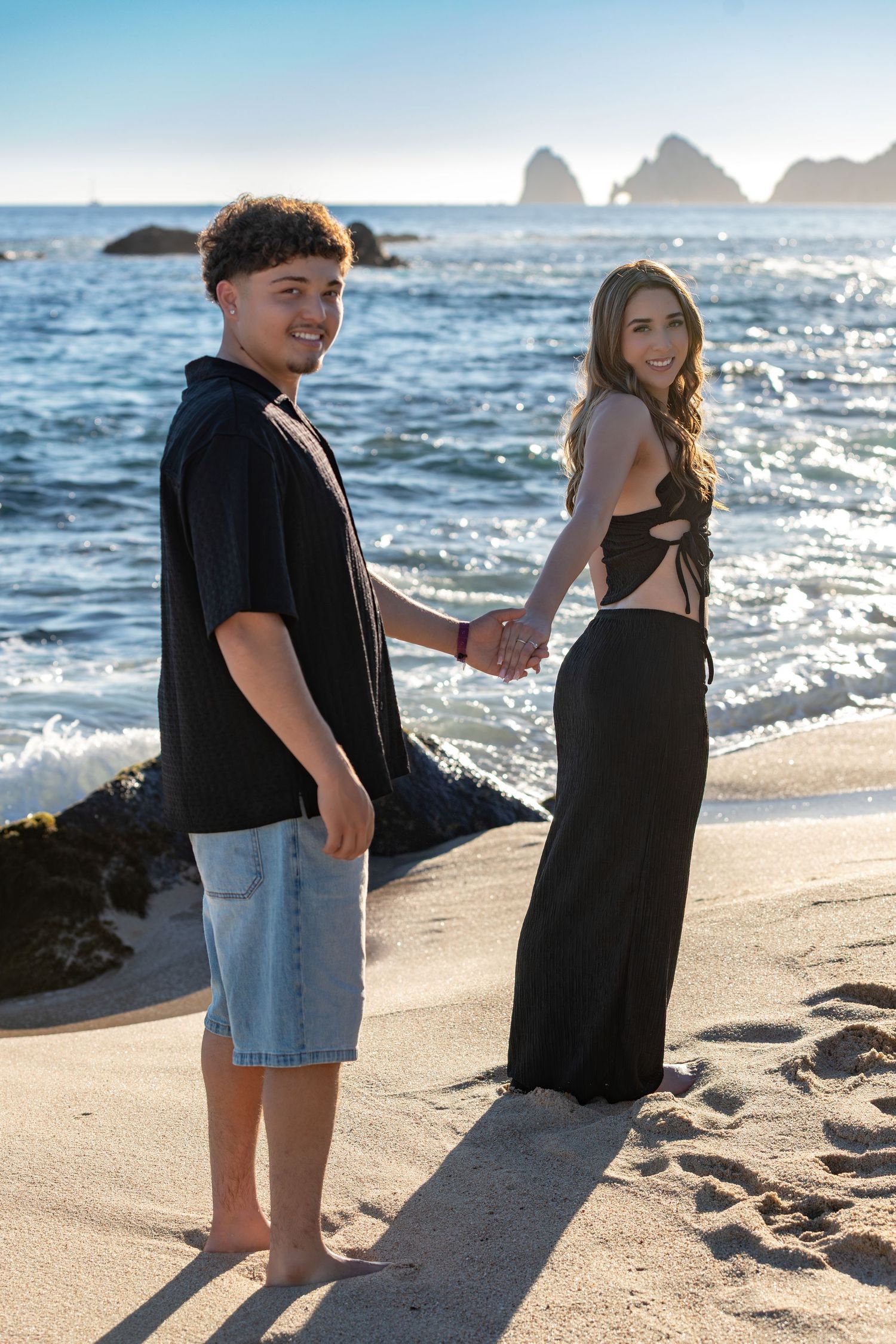 A couple holds hands on a sunny beach with ocean waves and rocky formations visible in the background.