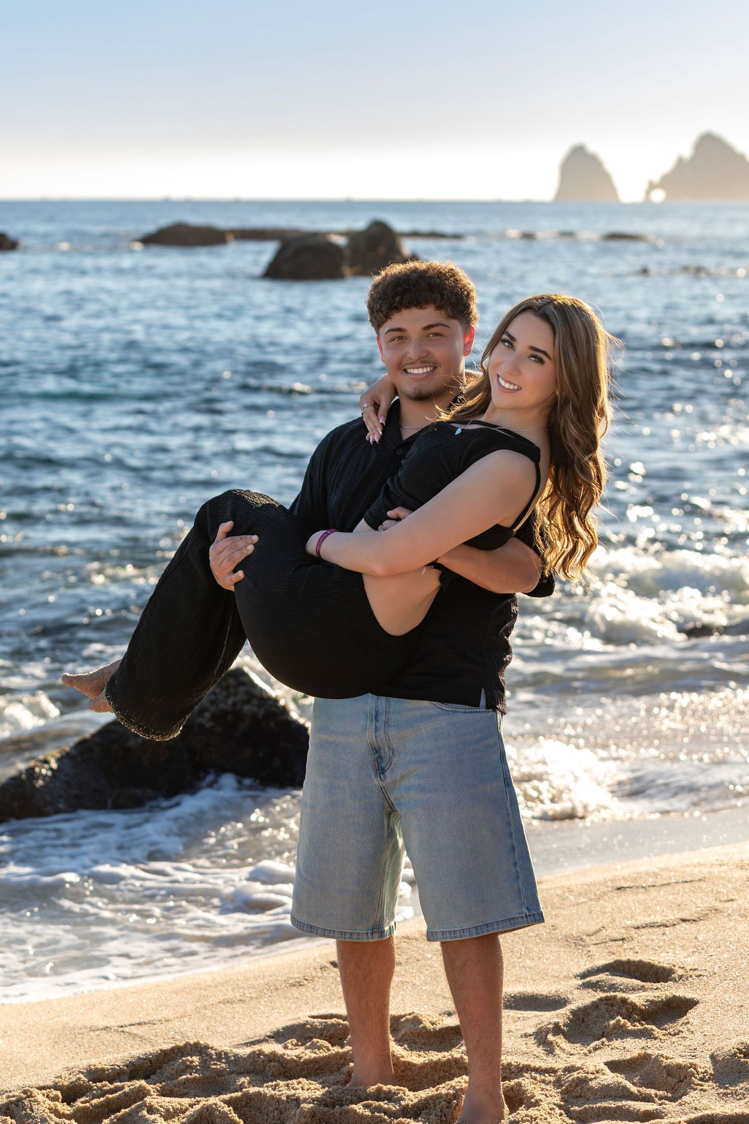 Playful couple embracing on a beach with rocky coastline and ocean waves in the background.