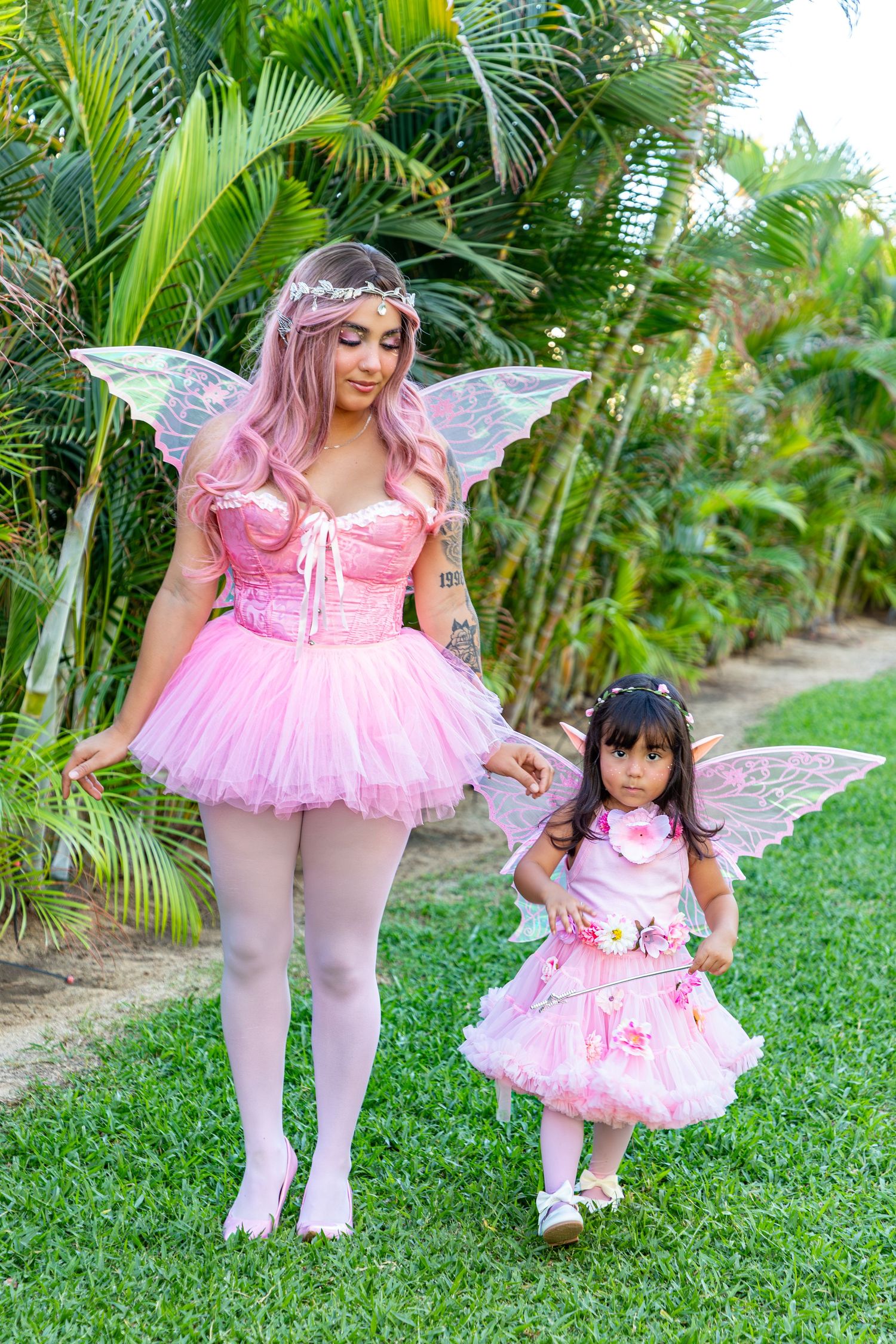 Pink fairy costumes with wings and tutus are showcased against a lush tropical palm garden backdrop.