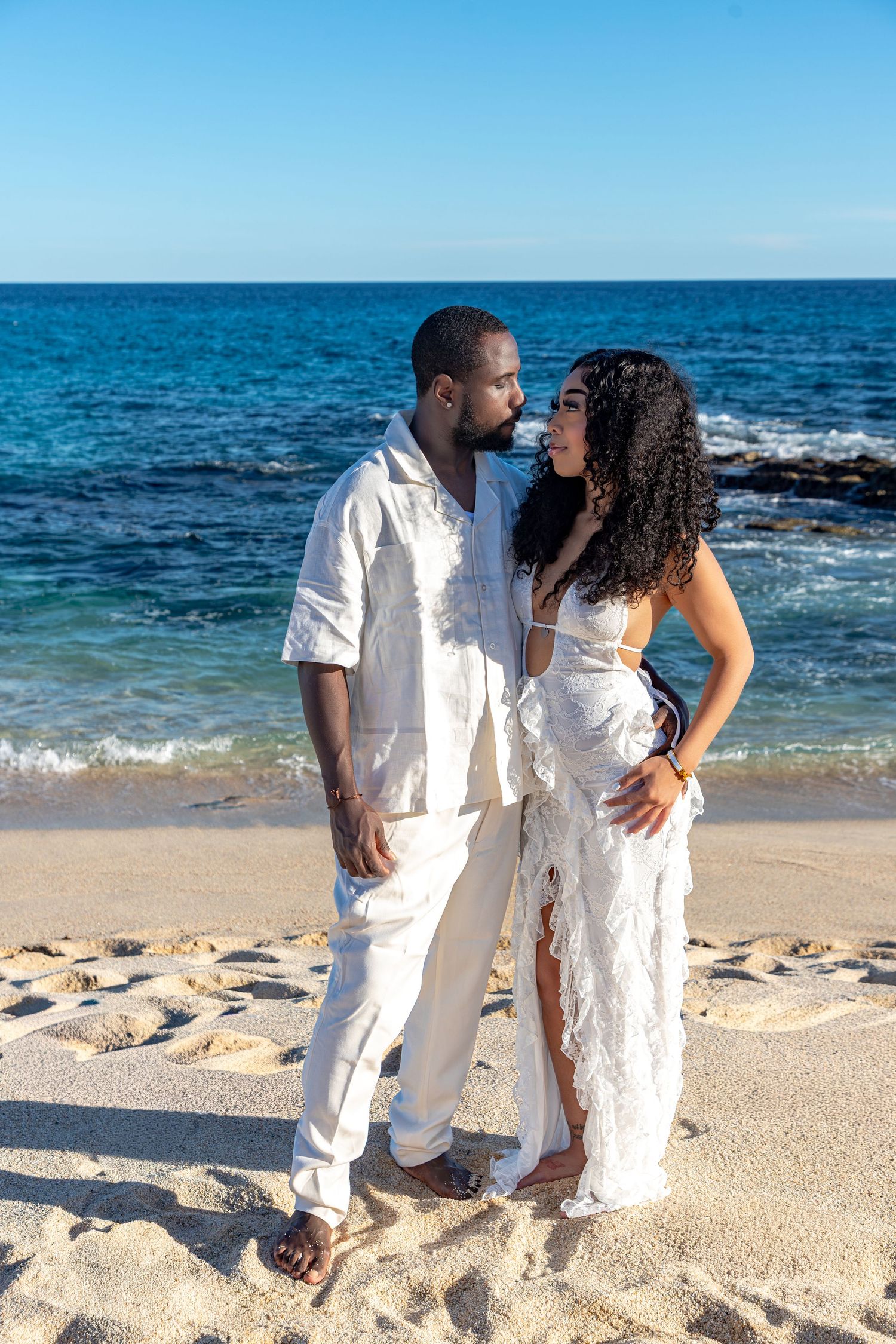 A romantic couple in white outfits shares intimate moments on a beautiful tropical beach with turquoise ocean waves.