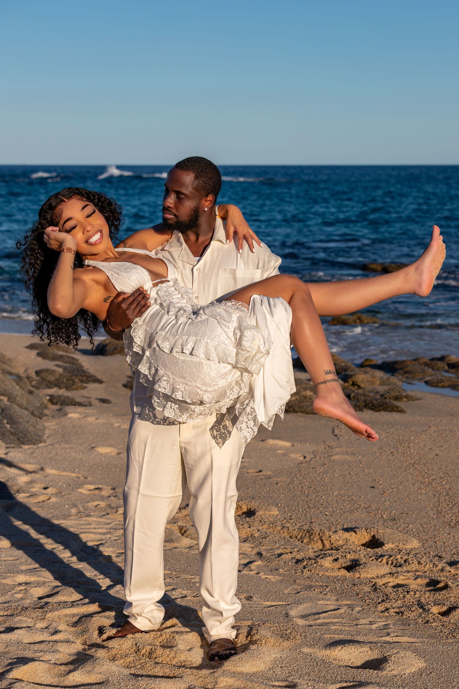 A couple in white outfits performs romantic dance moves on a sandy beach with blue ocean waters in the background.