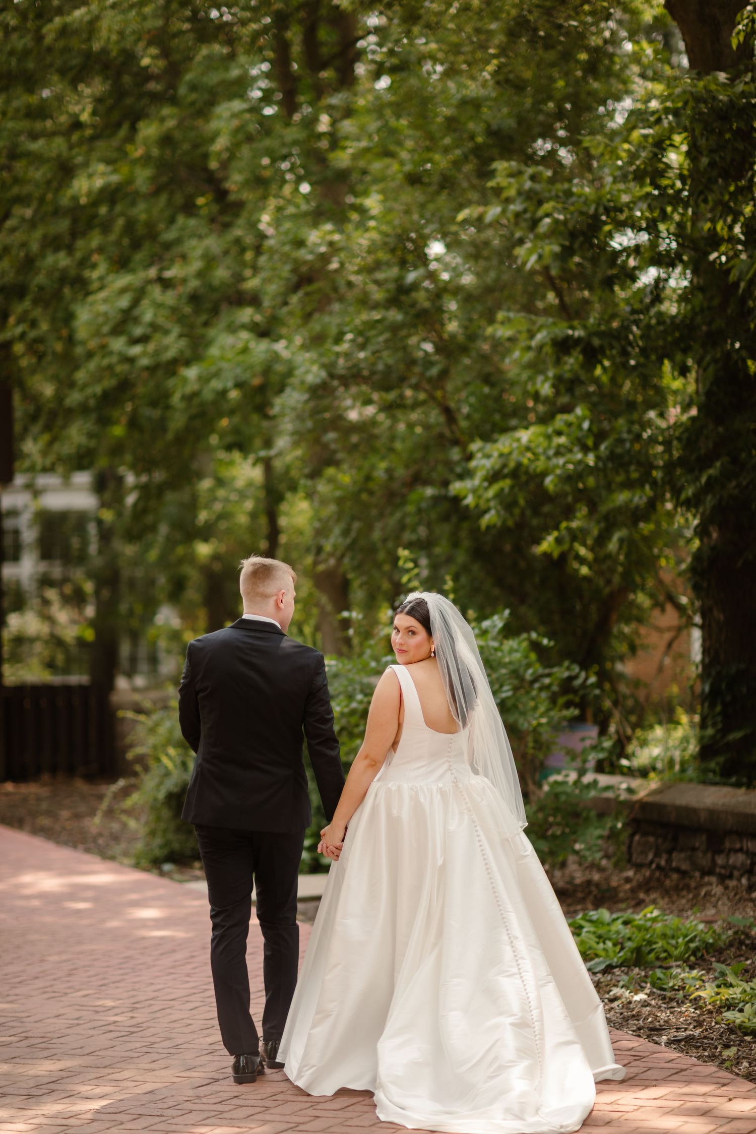 A bride and groom walk hand in hand down a tree-lined brick path during their wedding day.