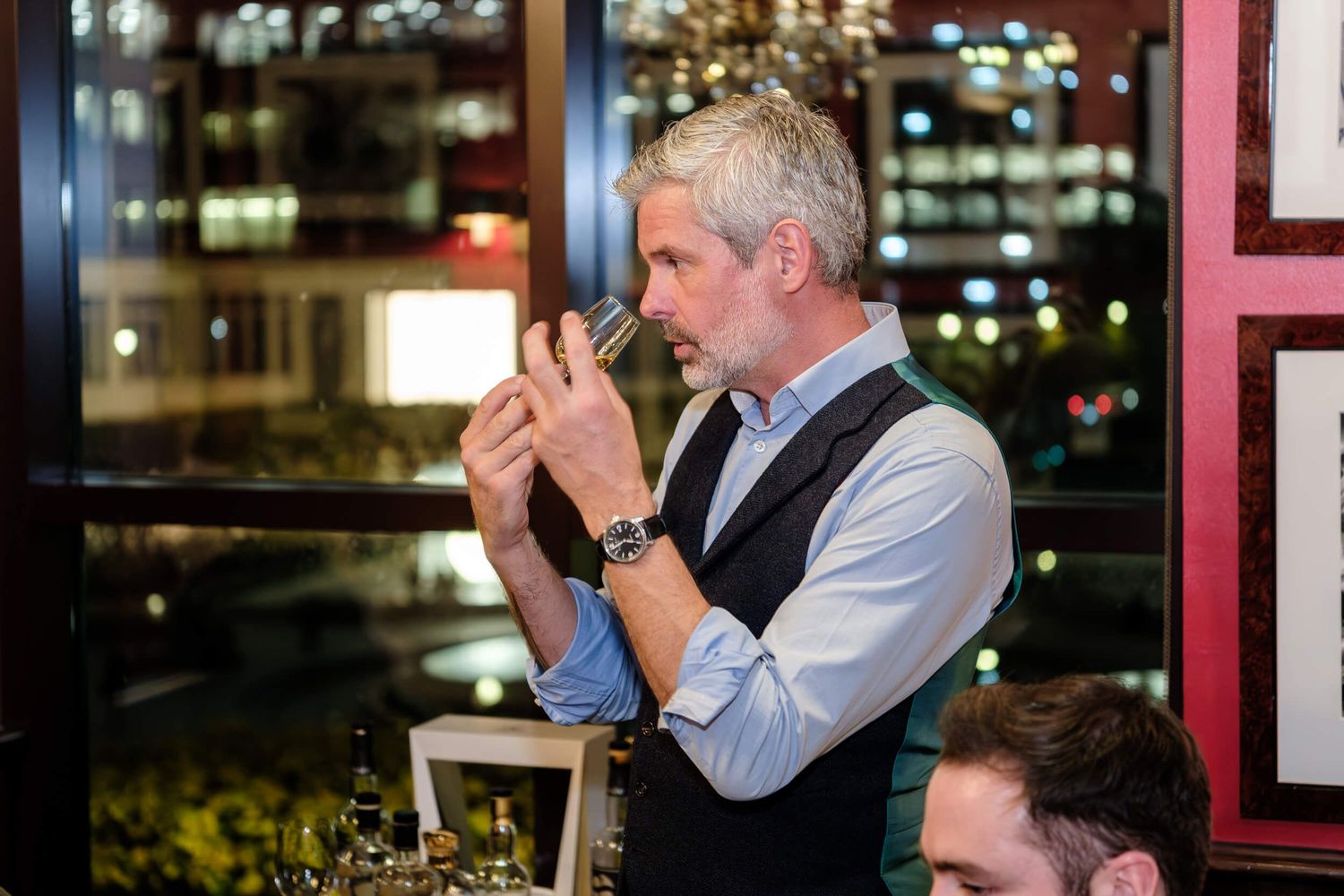 A server in formal attire lights a cigarette at a dimly lit restaurant with city lights visible in the background.