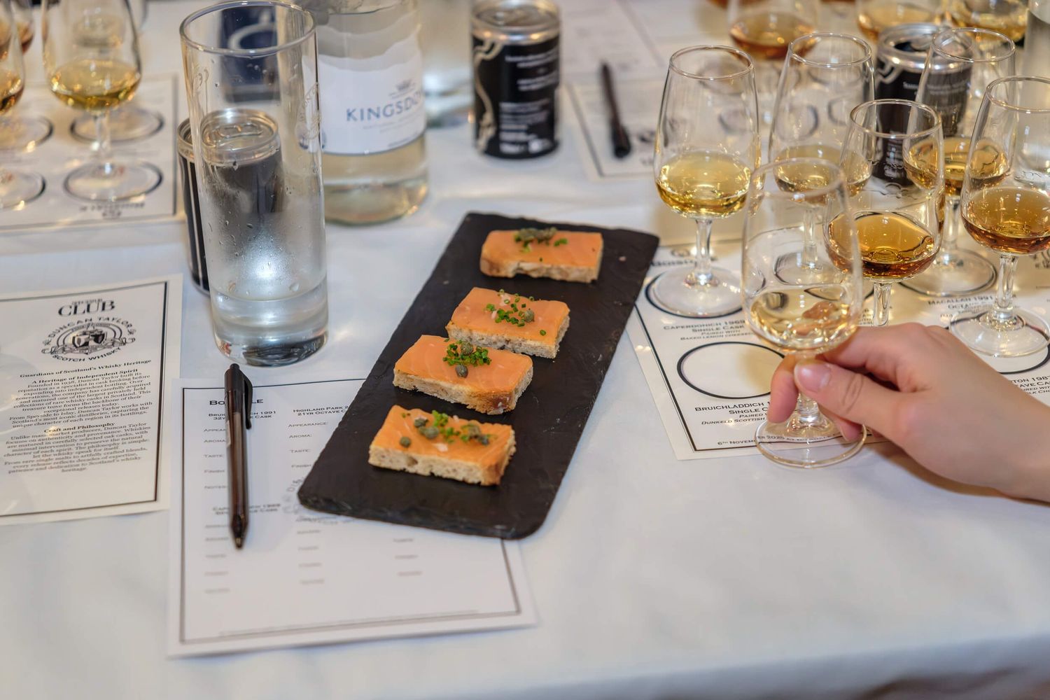 Several glasses of whiskey and appetizer plates are arranged on a white tablecloth during a tasting event.