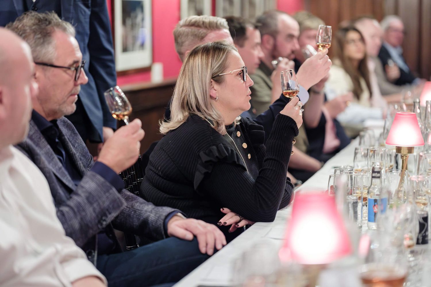 People seated at a long table participate in a wine tasting event with glasses raised and pink ambient lighting.