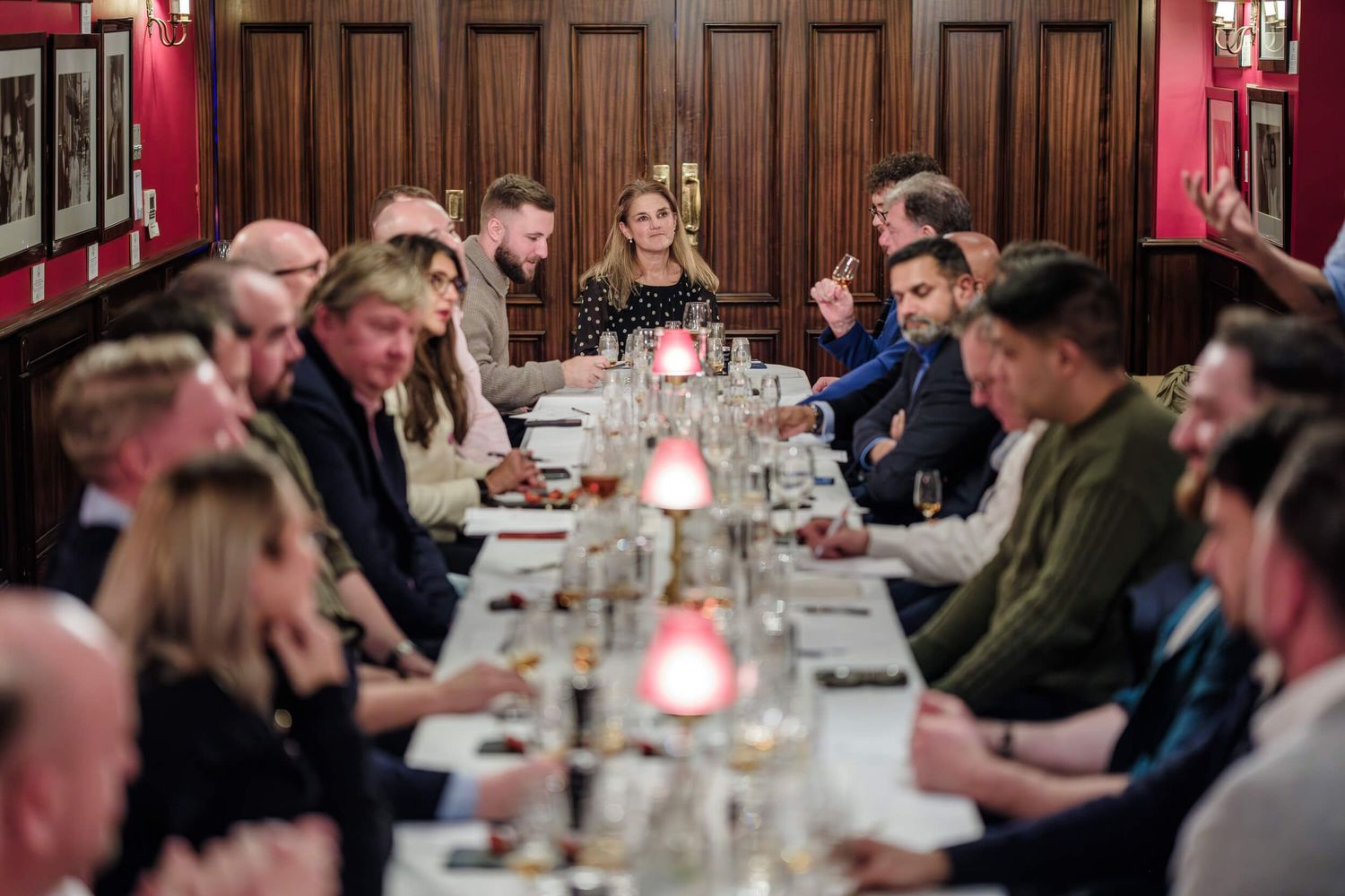 Guests gather around a long formal dining table with candlelight in an elegant wood-paneled room.