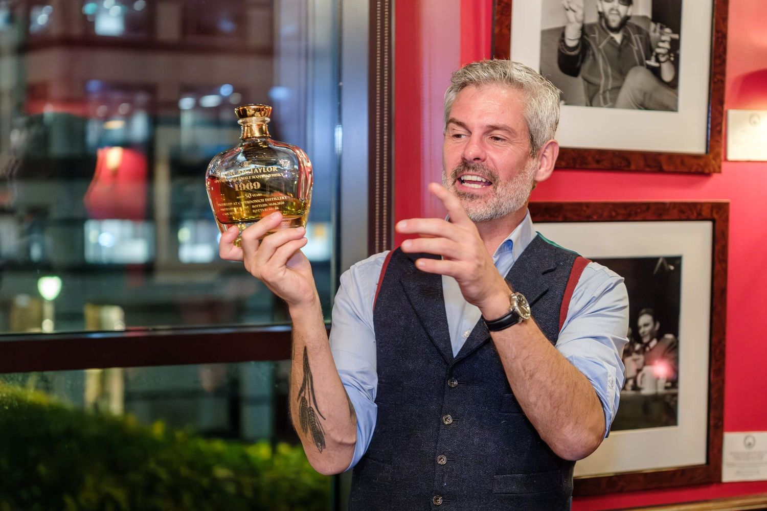A person in a vest holds up a bottle of whiskey while standing in front of framed photographs on a red wall.