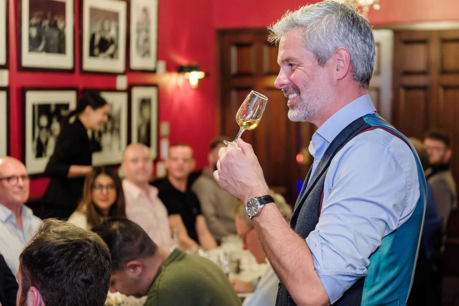 People gathered at a formal dinner event raise their wine glasses in a toast against a red wall backdrop.