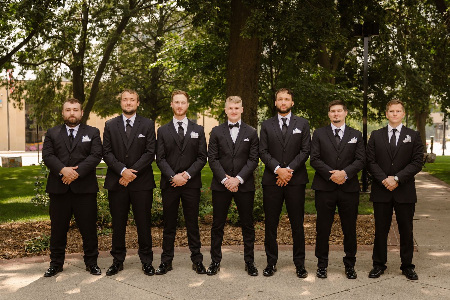 A row of groomsmen in black tuxedos and bow ties pose together outdoors in a formal wedding photo.