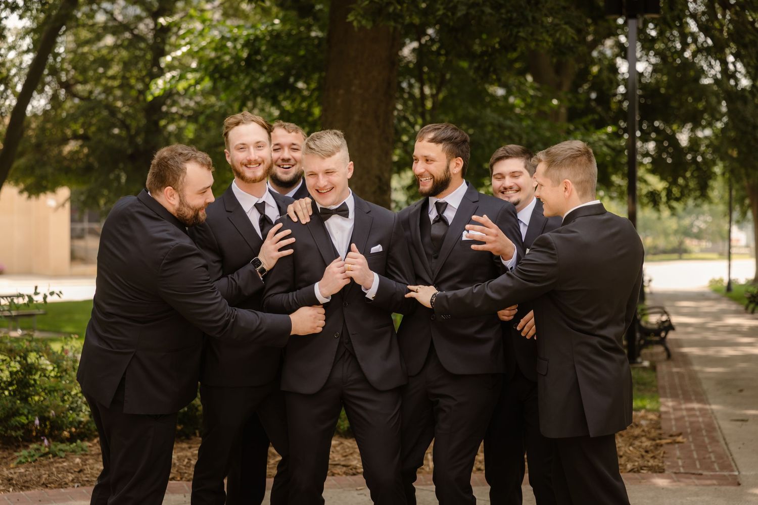 Groomsmen in black suits share laughs and drinks together during outdoor wedding celebration.
