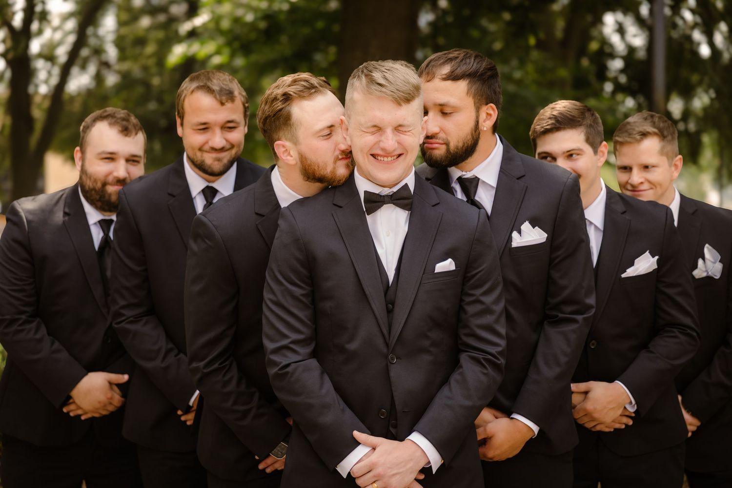 A group of groomsmen in black suits laugh together during a wedding photo session outdoors.