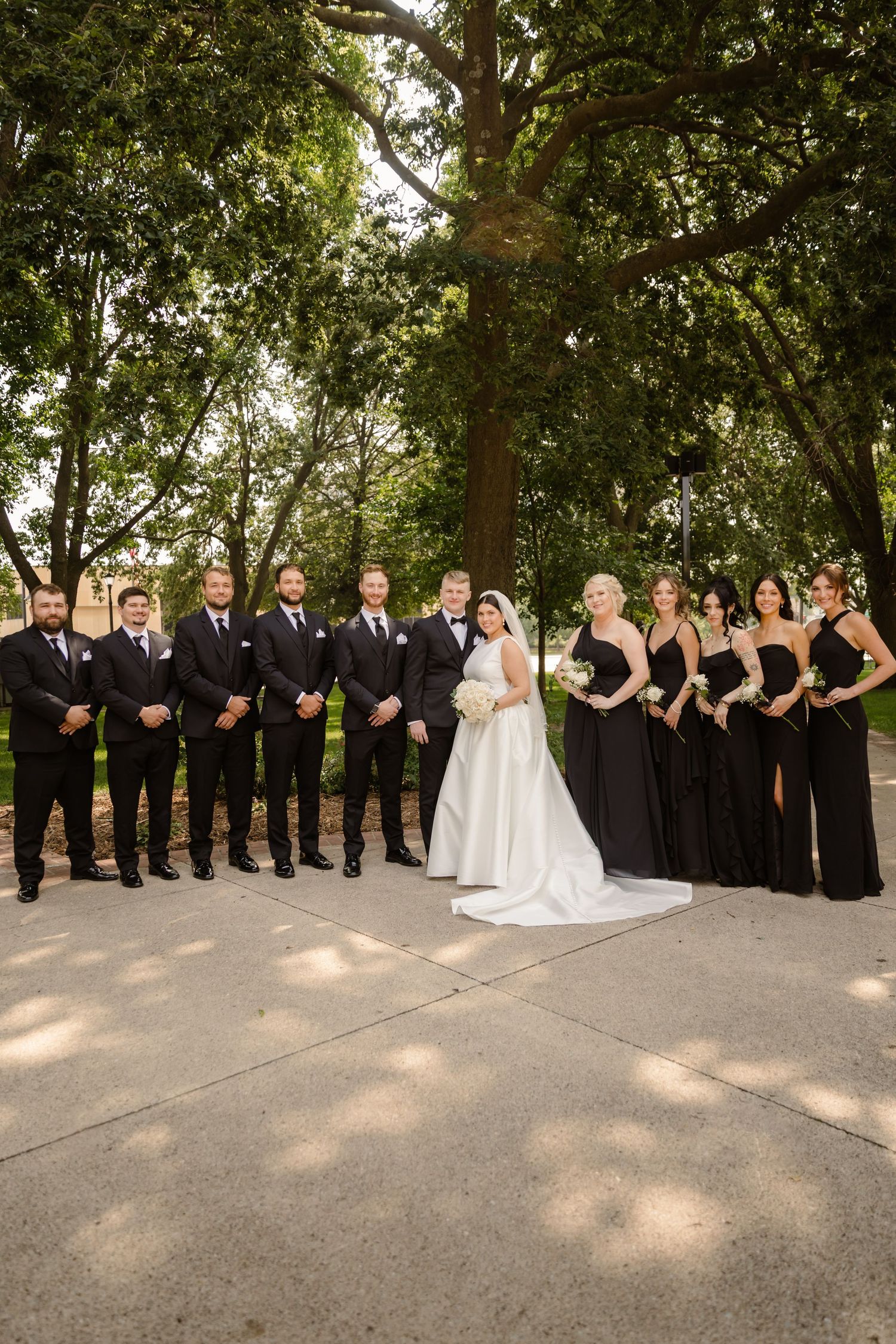 Wedding party in black and white attire poses in a line formation on a paved outdoor path surrounded by trees.