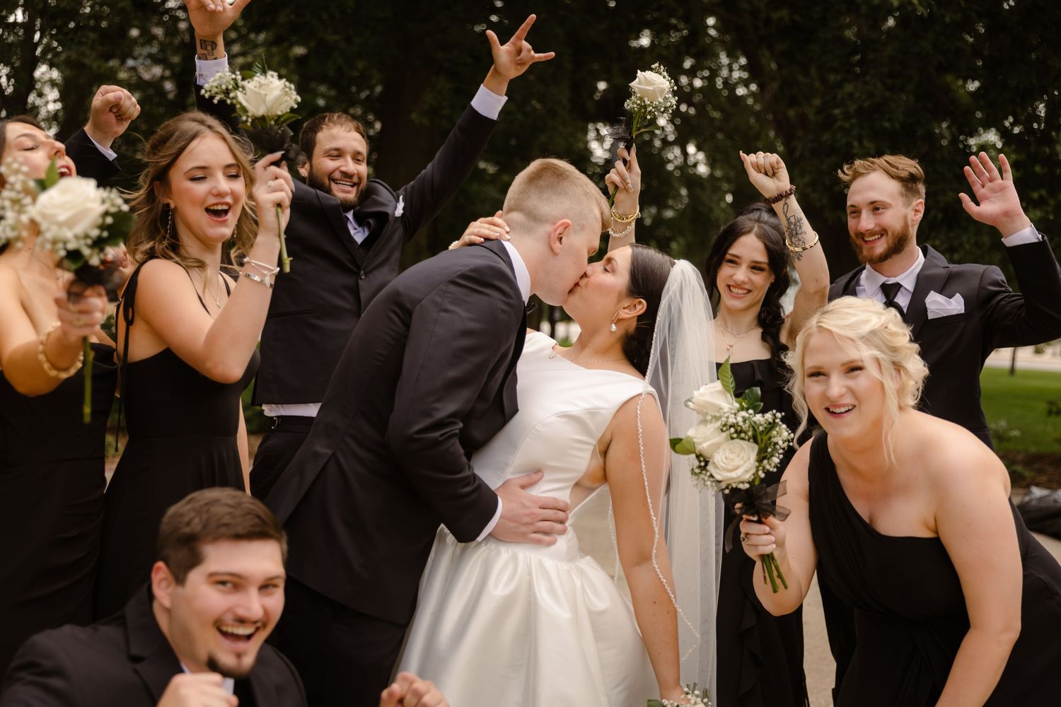 A joyful wedding party celebrates outdoors with raised hands and big smiles during a candid moment.