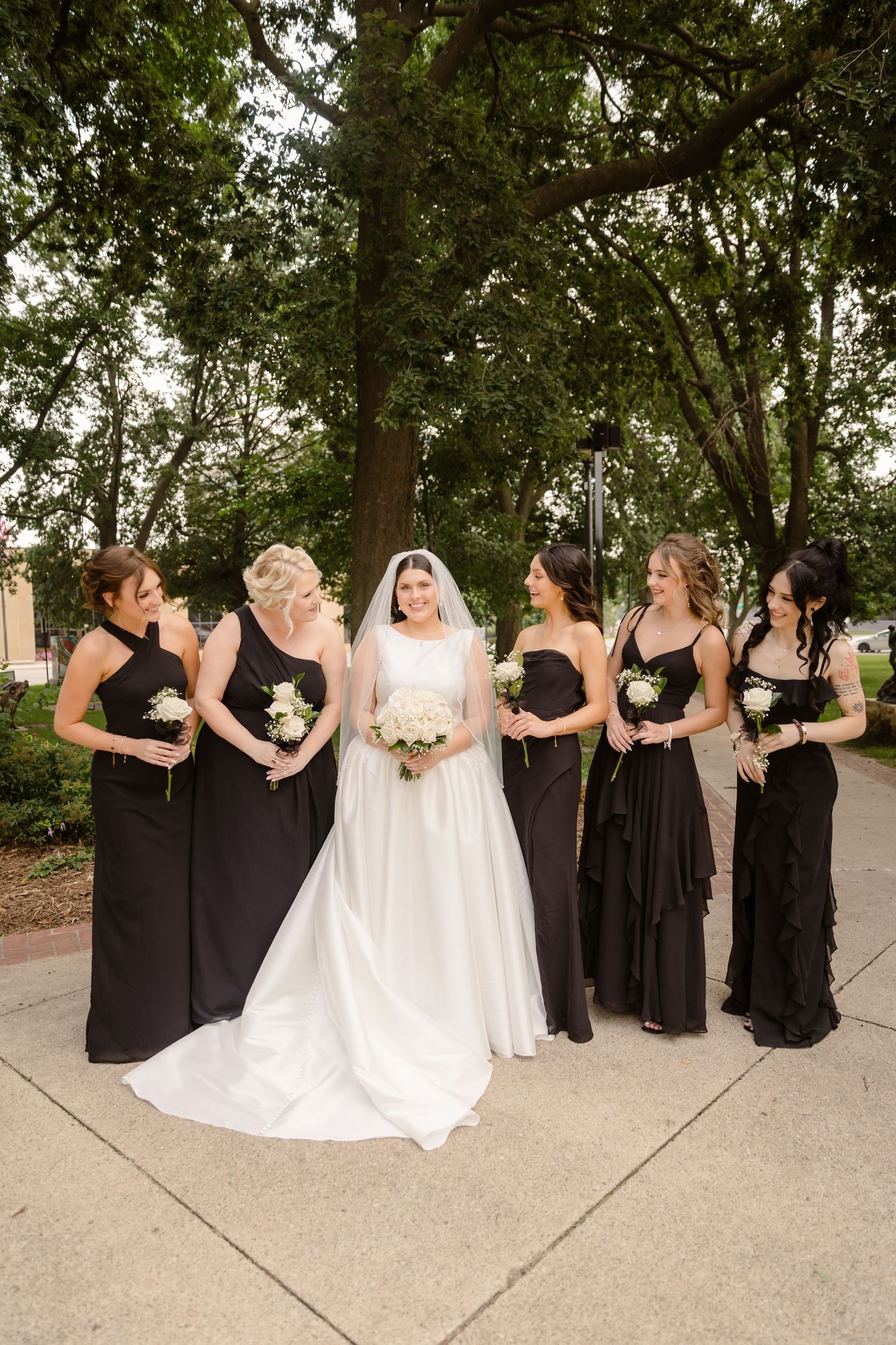 Wedding party in black bridesmaid dresses and white bridal gown pose together on outdoor pathway.