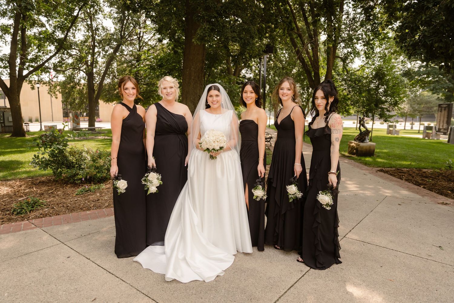 A wedding party in black and white attire poses outdoors on a tree-lined walkway on a sunny afternoon.