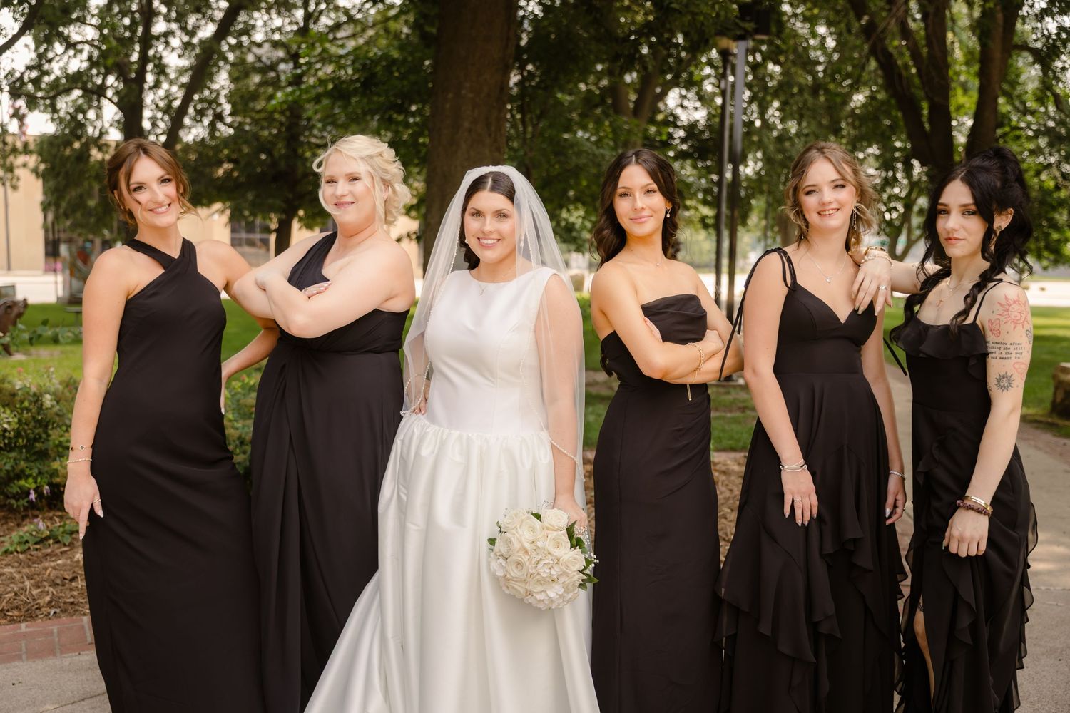 Wedding party poses together outdoors wearing black bridesmaid dresses and white bridal gown.