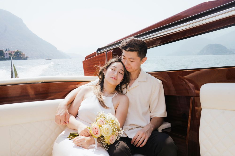 A couple relaxes on a luxury wooden boat with mountains and water in the background, holding a floral bouquet.