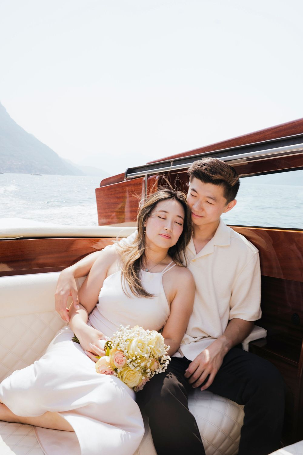 Romantic couple lounging together on a vintage wooden boat with yellow flowers in a dreamy coastal setting.
