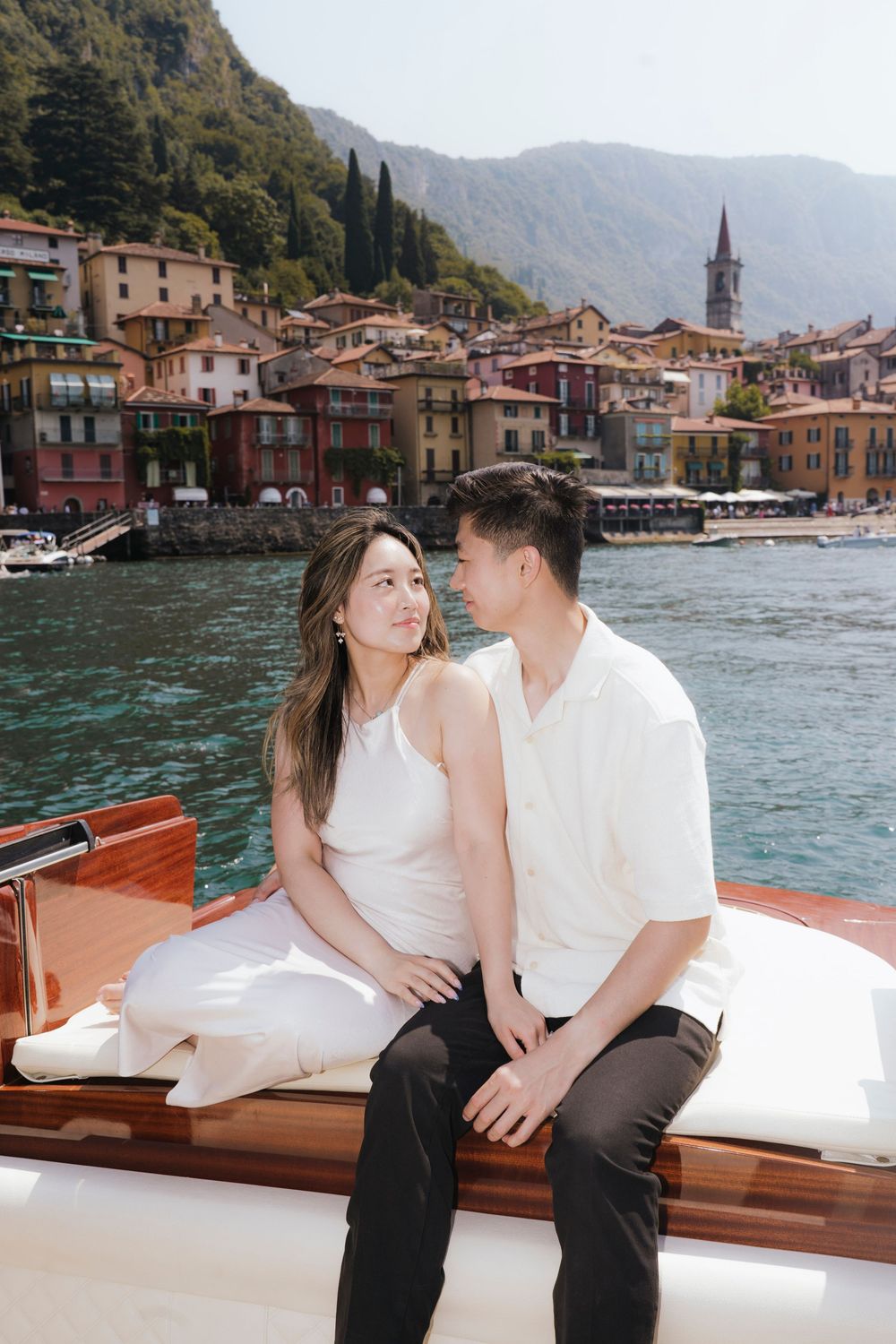 Couple enjoying an intimate moment on a classic boat while cruising past colorful Italian lakeside buildings.