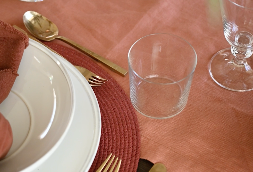 Elegant table setting with white plates, gold utensils, and wine glasses on a coral-colored tablecloth.