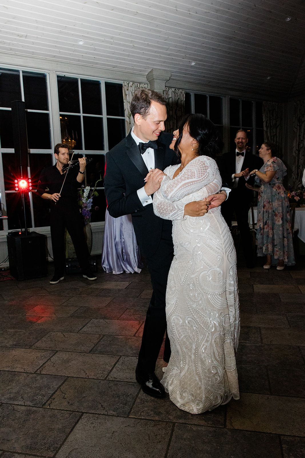 A couple shares their first dance at their wedding reception on a stone patio while guests watch in the background.
