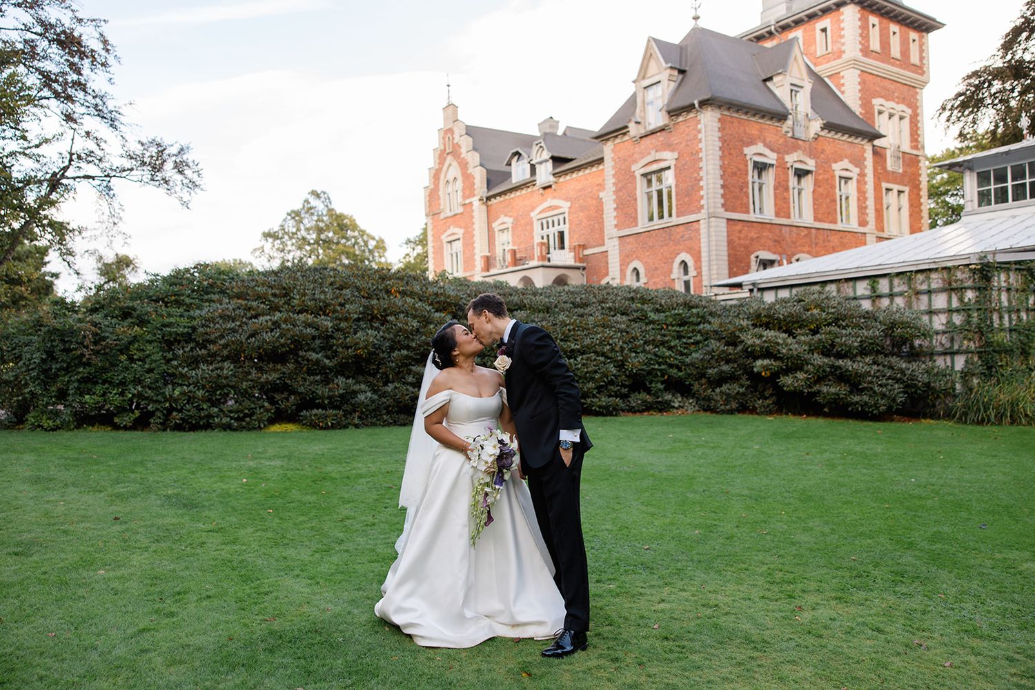 A couple in wedding attire embraces on a lawn in front of a grand Victorian-style brick mansion.